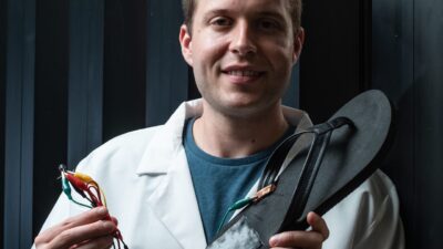 Rice University postdoctoral researcher Michael Stanford holds a flip-flop with a triboelectric nanogenerator, based on laser-induced graphene, attached to the heel. Walking with the flip-flop generates electricity with repeated contact between the generator and the wearer’s skin. Stanford wired the device to store energy on a capacitor. Courtesy: Jeff Fitlow/Rice University