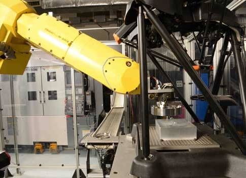 The robot places a machined bearing into the Equator’s measuring envelope (foreground), with part conveyor and Okuma 2SP-250H twin-spindle lathe in background. The aluminum block on the Equator’s fixture plate has a hole in the center, which the Equator u